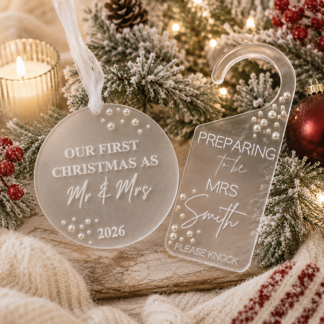 Two personalized frosted acrylic Christmas door hangers and round ornament displayed on a rustic wooden surface with holiday decorations, including pine branches, a lit candle, berries, and ornaments.