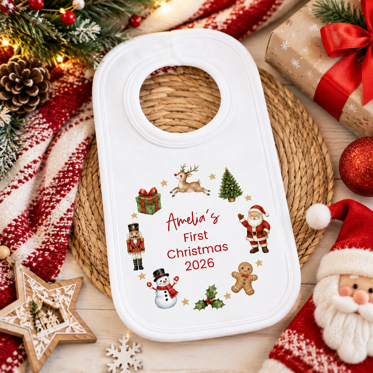 A white baby bib with a circular neck opening, decorated with Christmas illustrations and the personalized text 'Amelia's First Christmas 2026'. The bib is styled in a festive flat-lay setting with a red and white striped knit blanket, a woven placemat, wrapped gifts, a star ornament, and a Santa hat.