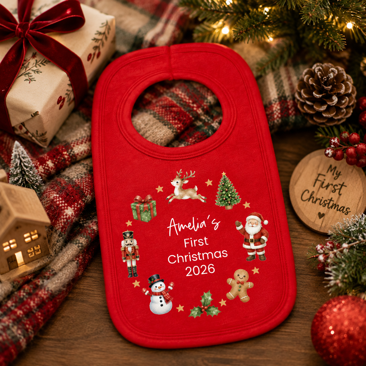A red baby bib with personalized Christmas text reading 'Amelia's First Christmas 2026' and festive embroidered illustrations, styled on a wooden surface with holiday decorations including a plaid blanket, wrapped gift, pinecones, evergreen branches, and a wooden sign.