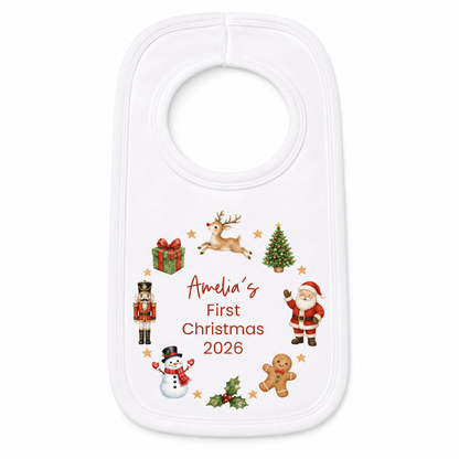A white baby bib with a personalized Christmas design, featuring illustrations of Santa Claus, a reindeer, a nutcracker, a snowman, a gingerbread man, a Christmas tree, and a gift box around the text 'Amelia's First Christmas 2026'.