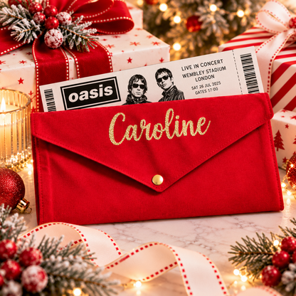 A red velvet envelope-style pouch with gold script reading 'Caroline' and a gold snap button, styled on a holiday table with gift boxes, ornaments, a concert ticket, and warm string lights.