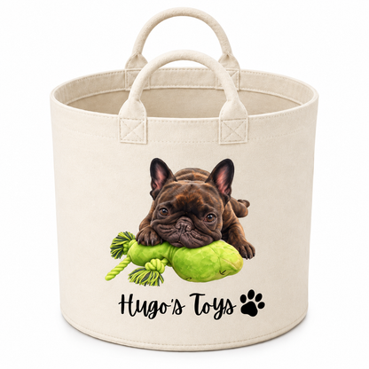 A cream-colored fabric pet toy basket featuring a printed illustration of a brown French Bulldog holding a green rope toy, with the text 'Hugo's Toys' and a paw print below.