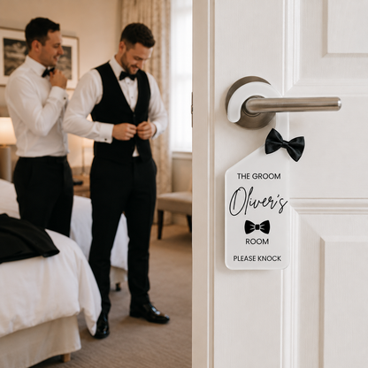 A white personalized room tag with a black bowtie graphic hangs on a white hotel door handle in a hotel room. In the background, two men in white shirts and black bowties are adjusting their attire in a well-lit bedroom.