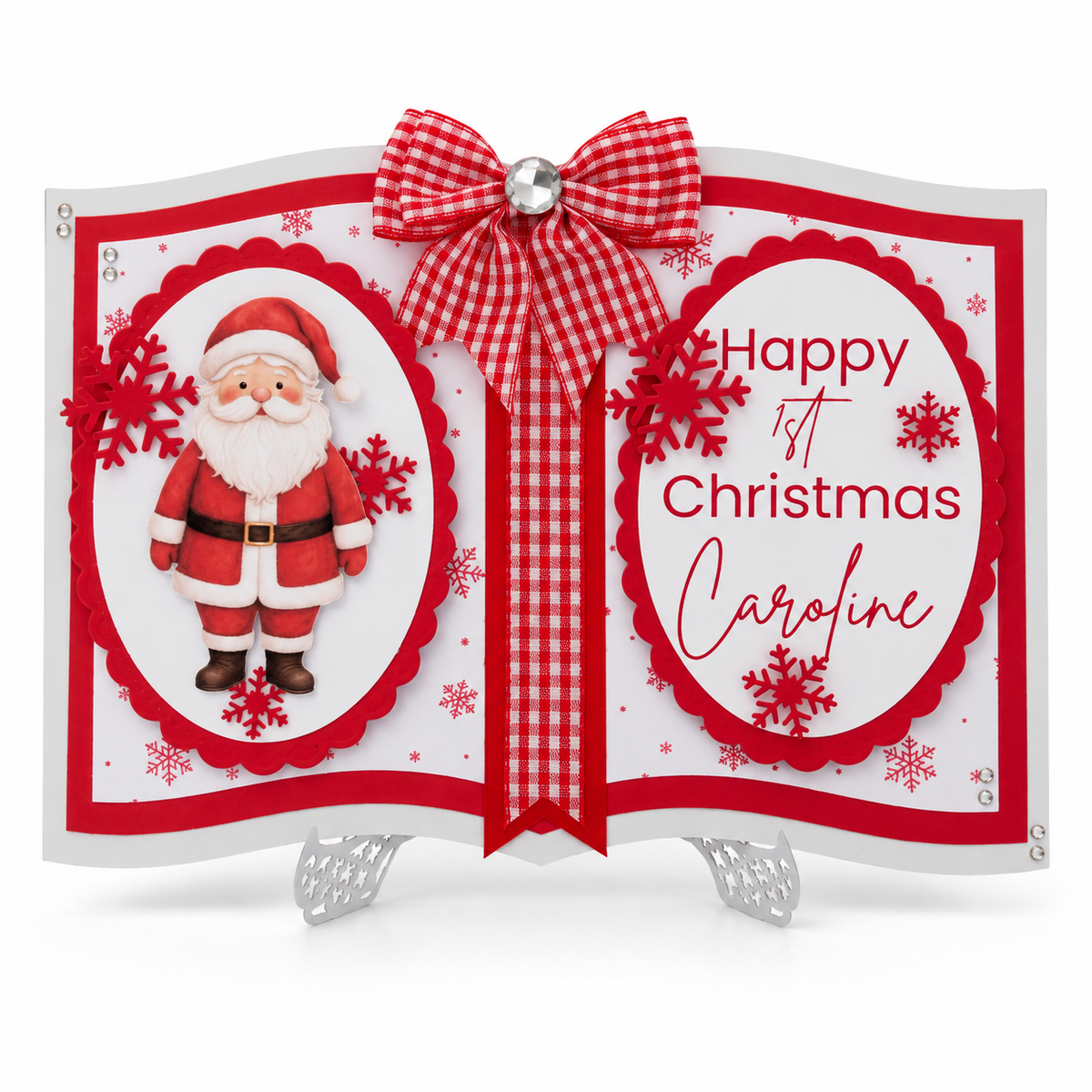 A festive first Christmas card displayed on a small silver stand. The card is designed like an open book with red and white gingham ribbon accents, a red bow, a photo of Santa Claus on the left page, and the text 'Happy 1st Christmas Caroline' on the right page, decorated with red snowflakes and white snowflakes.