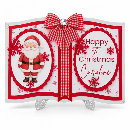 A festive first Christmas card displayed on a small silver stand. The card is designed like an open book with red and white gingham ribbon accents, a red bow, a photo of Santa Claus on the left page, and the text 'Happy 1st Christmas Caroline' on the right page, decorated with red snowflakes and white snowflakes.