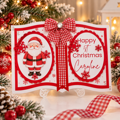 A festive first birthday Christmas pop-up card displayed on a small white easel. The card is designed like an open book with red and white gingham ribbon accents, a red bow, and a 3D Santa Claus figure on the left page.
