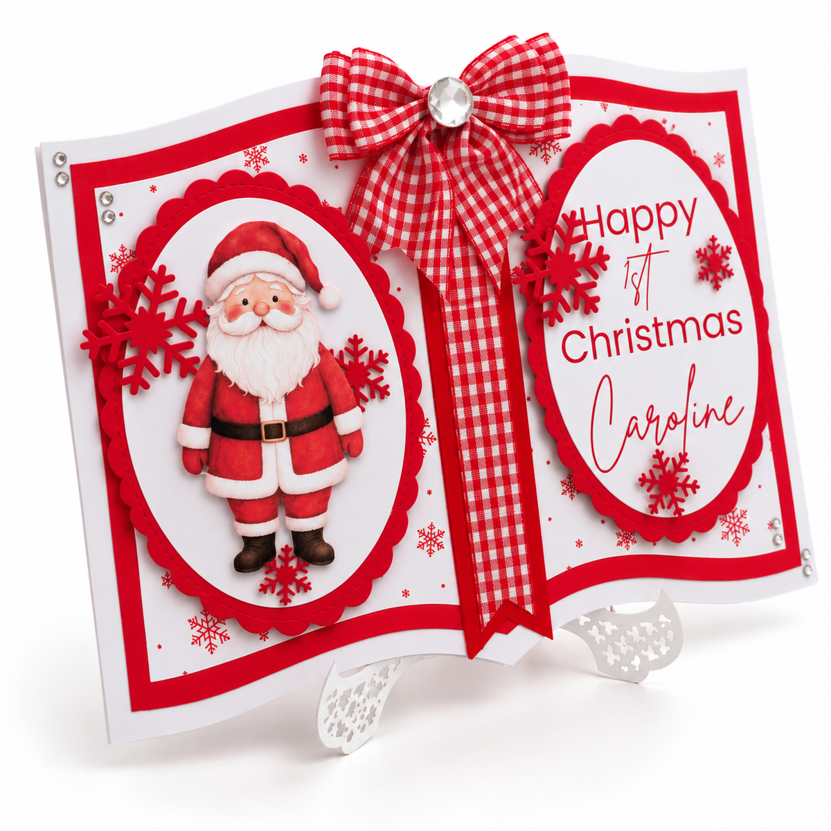 A handmade Christmas greeting card displayed on a small white stand. The card is shaped like an open book with red and white borders, a red-and-white gingham bow at the top, a 3D Santa Claus figure on the left page, and the text 'Happy 1st Christmas Caroline' on the right page.