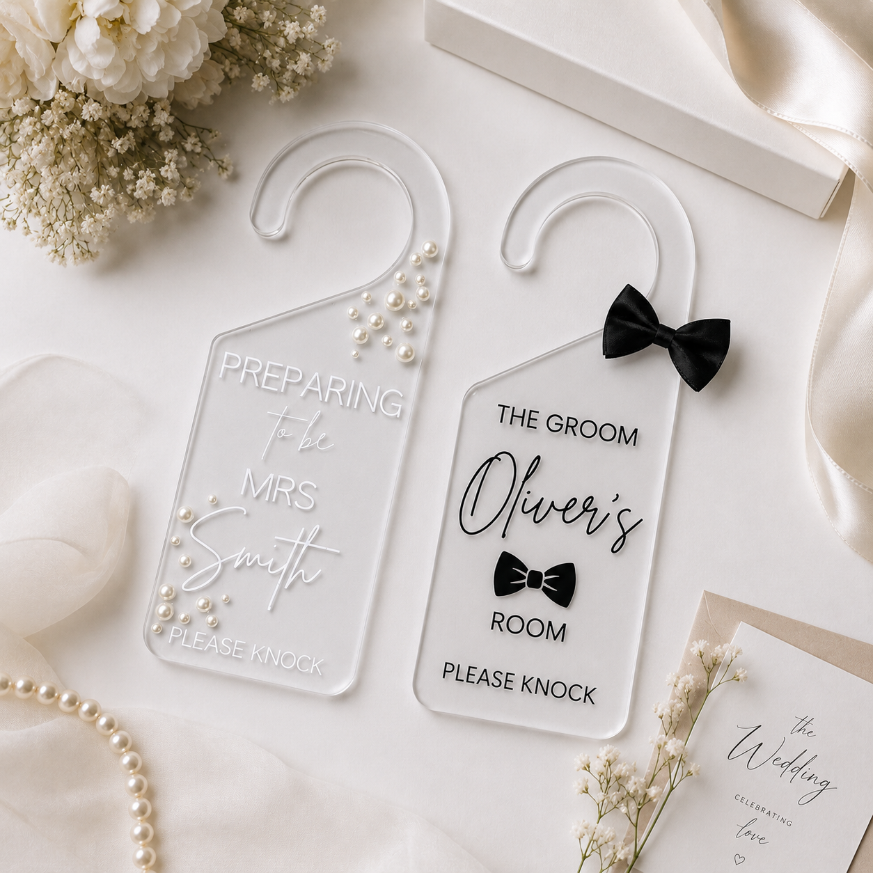Two clear acrylic hanger-style door hoods with wedding-themed printed text, arranged on a soft white surface with flowers, pearls, and a ribbon. One reads 'Preparing to be Mrs.