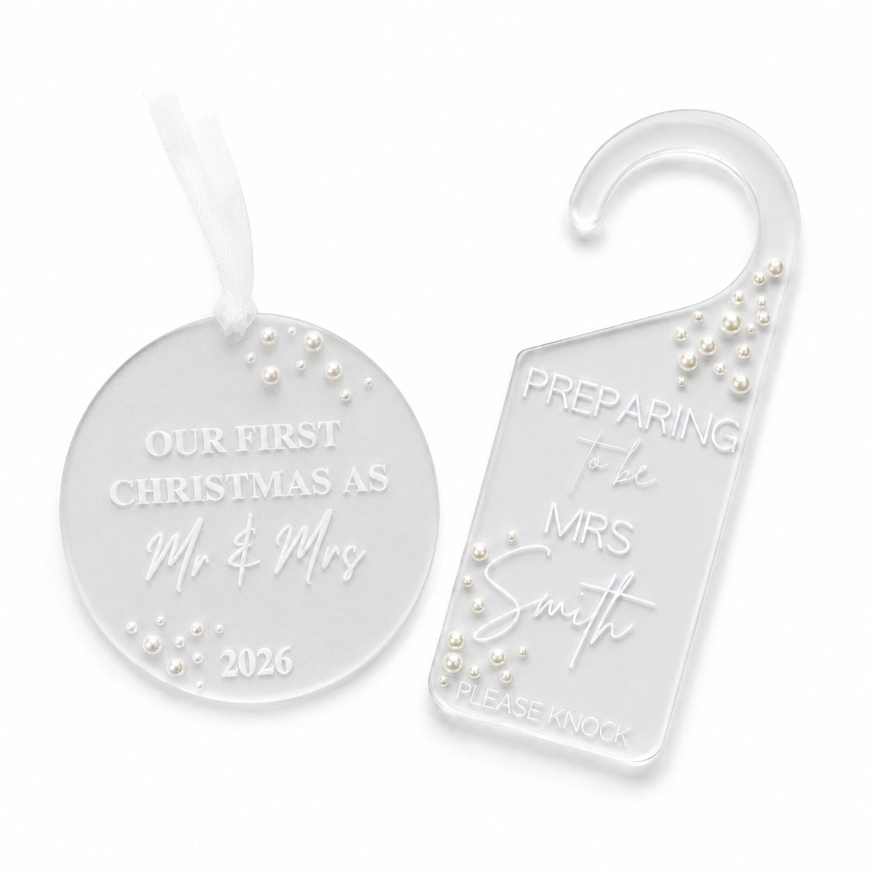 Two clear acrylic holiday keepsakes on a white background: a round ornament with a white ribbon and a house hook tag. Both are engraved with personalized text and decorated with small white pearl-like beads.
