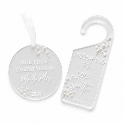Two clear acrylic holiday keepsakes on a white background: a round ornament with a white ribbon and a house hook tag. Both are engraved with personalized text and decorated with small white pearl-like beads.