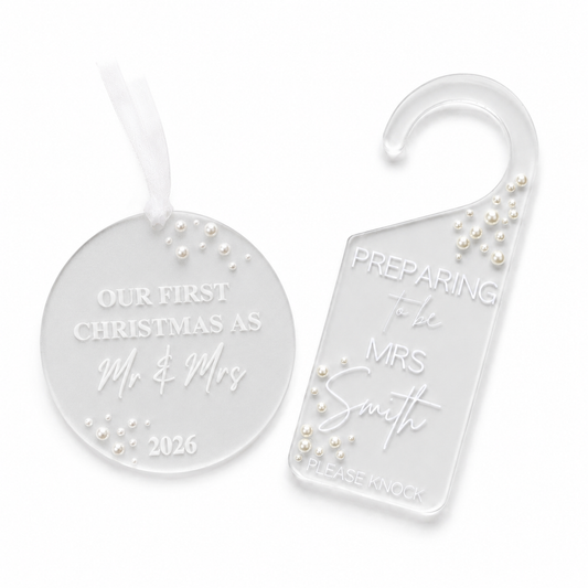 Two clear acrylic holiday keepsakes on a white background: a round ornament with a white ribbon and a house hook tag. Both are engraved with personalized text and decorated with small white pearl-like beads.