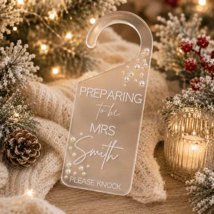 A clear acrylic hanging door hanger with the text 'PREPARING to be MRS Smith PLEASE KNOCK' and decorative pearl-like dots, styled on a knit blanket with holiday greenery, pinecones, and warm string lights.