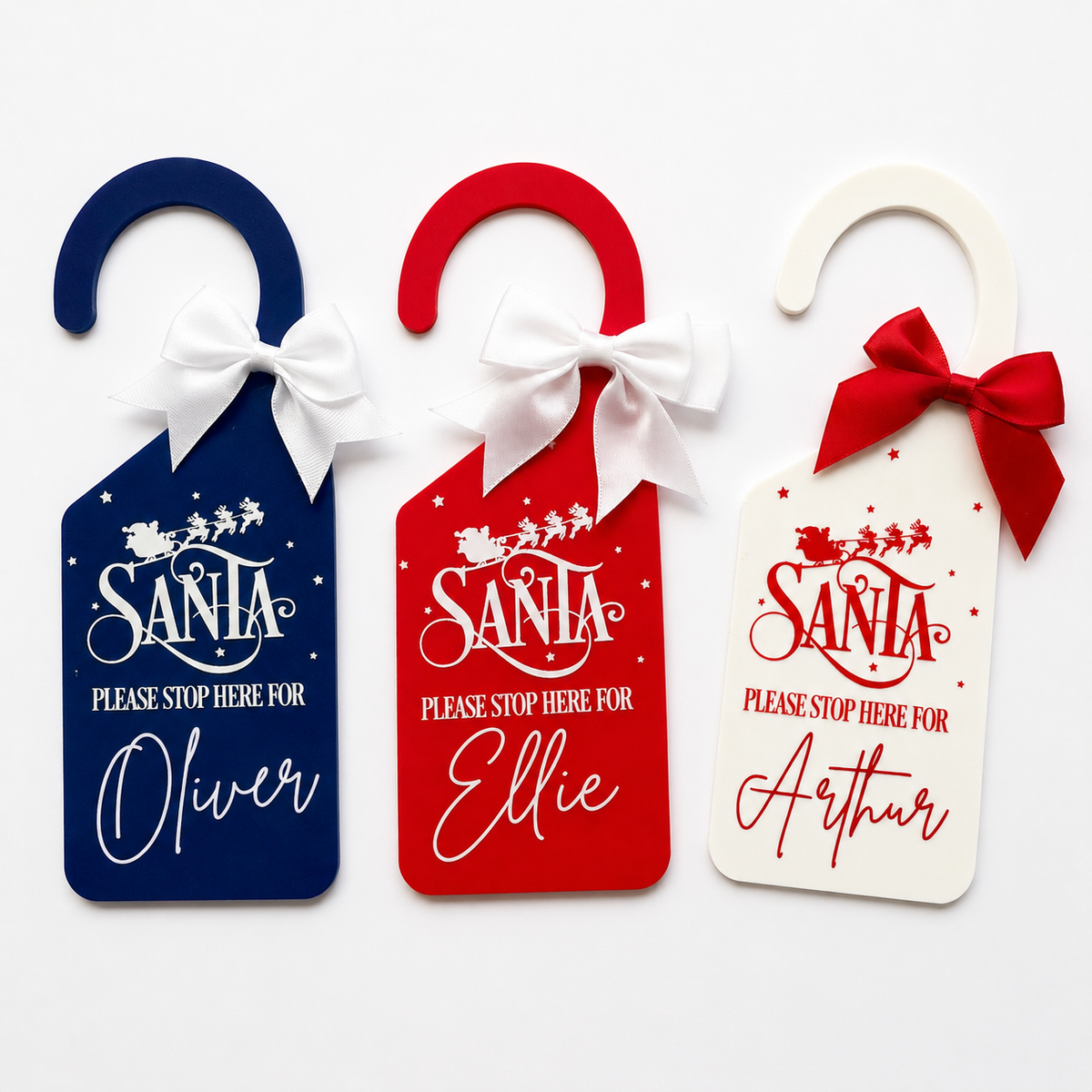 Three personalized Christmas Santa door hangers in navy blue, red, and white, each with a white bow and a custom name printed below the text 'Please Stop Here For Santa'.