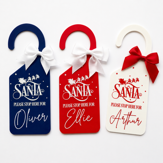 Three personalized Christmas Santa door hangers in navy blue, red, and white, each with a white bow and a custom name printed below the text 'Please Stop Here For Santa'.