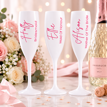 Three white champagne flutes with personalized names in pink script and black serif text, displayed on a marble surface with pink roses, a Prosecco bottle, and confetti in the background.