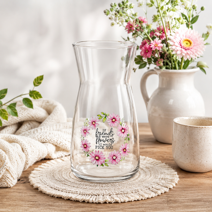 Clear glass pitcher with floral design and text on a wooden surface with flowers and a mug in the background.