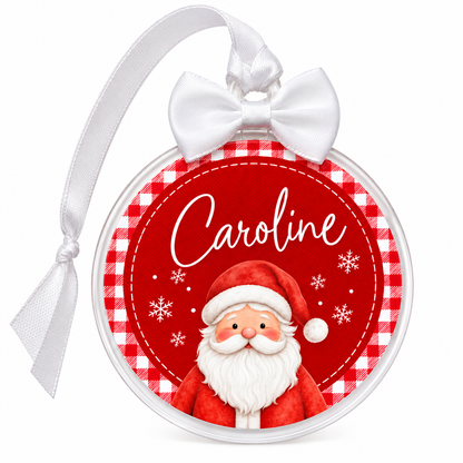 A personalized Christmas ornament featuring a red and white gingham border, a red background with white snowflakes, the name 'Caroline' in white script, and a watercolor-style Santa Claus illustration. The ornament has a white satin bow and hanging ribbon on a plain white background.