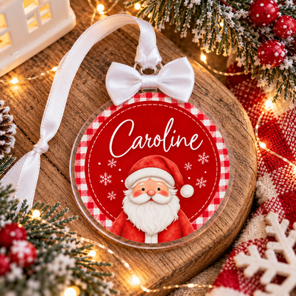 A personalized Christmas ornament featuring a cartoon Santa Claus on a red and white gingham background with the name 'Caroline' in white script. The ornament is decorated with a white bow and ribbon, displayed on a wooden surface surrounded by festive holiday decorations, including pine branches, red berries, and warm string lights.