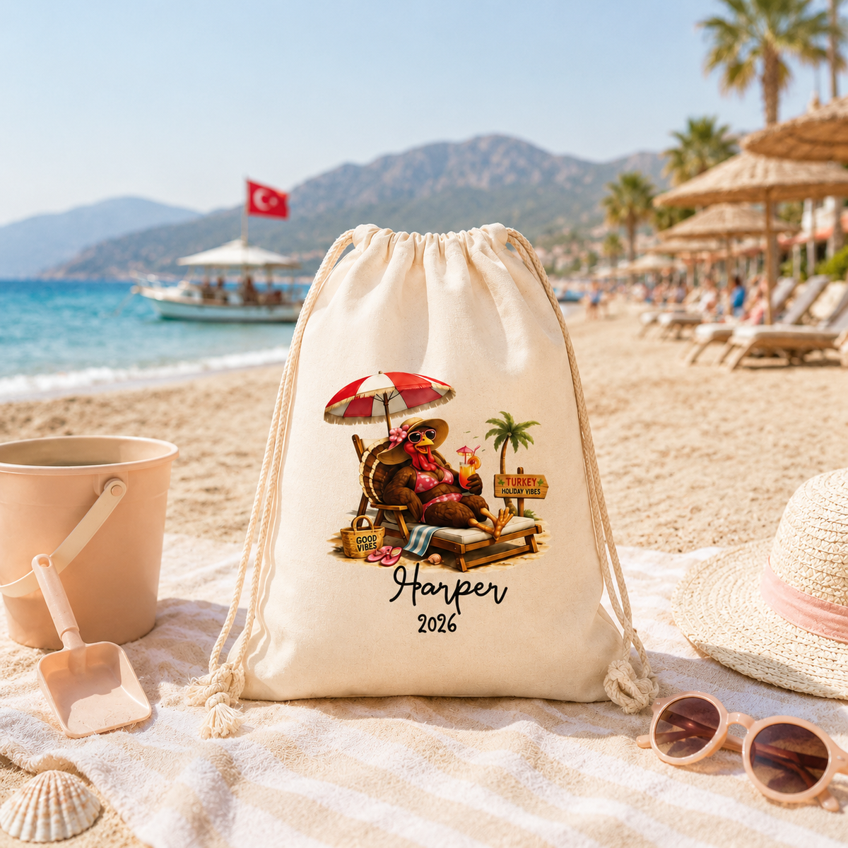 A cream-colored drawstring beach bag with a custom cartoon illustration and the text 'Harper 2026' is displayed on a beach blanket with beach-themed props like a straw hat, sunglasses, a sand bucket, and a shovel. The background shows a sunny beach scene with the blue ocean, mountains, and palm trees.