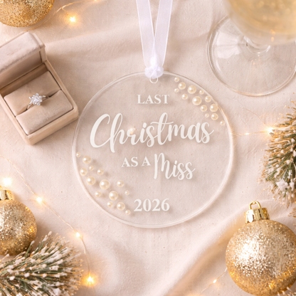 Clear ornament with 'Last Christmas as a Miss 2026' text, surrounded by festive decorations on a light background.