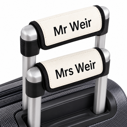 Custom luggage handle covers reading 'Mr Weir' and 'Mrs Weir' in black text on an off-white background, shown on silver telescoping handles of a suitcase.