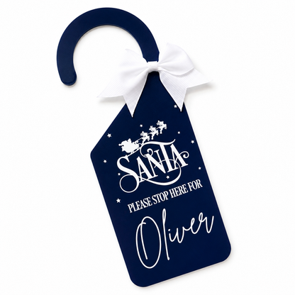 A navy blue Christmas door hanger with a white bow at the top. The tag features white printed text reading 'Santa please stop here for Oliver' with Santa and sleigh graphics above, shown on a plain white background.