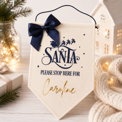 Decorative sign with 'Santa Please Stop Here For Caroline' text, navy bow, and Christmas-themed background.