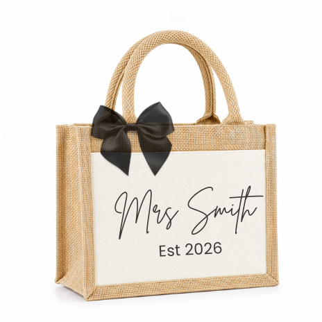 A personalized jute gift bag with a cream fabric front panel, black script text reading 'Mrs Smith Est 2026', and a black satin bow on the handle, shown on a white background.