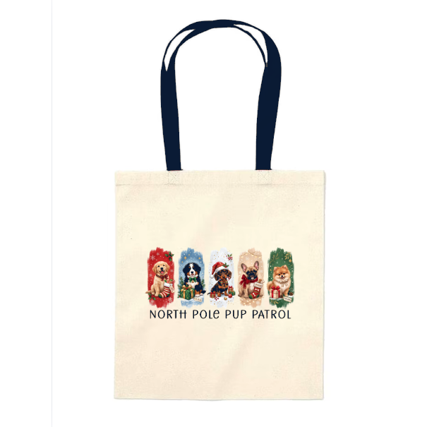 An off-white canvas tote bag with dark navy blue handles, featuring a graphic of four cute cartoon dogs dressed in winter hats and scarves under the text 'NORTH POLE PUP PATROL'.