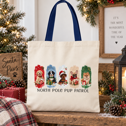 A cream canvas tote bag with dark blue handles, decorated with a Christmas-themed graphic of four dog breeds in Santa hats and the text 'NORTH POLE PUP PATROL'. The bag is displayed on a wooden shelf surrounded by holiday decorations including a Christmas tree, pinecones, a lantern, and a plaid blanket.