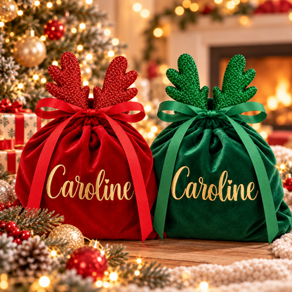 Two personalized Christmas gift bags in red and green velvet with gold 'Caroline' script, topped with glittery antler bows, displayed on a holiday table with warm lights and ornaments.