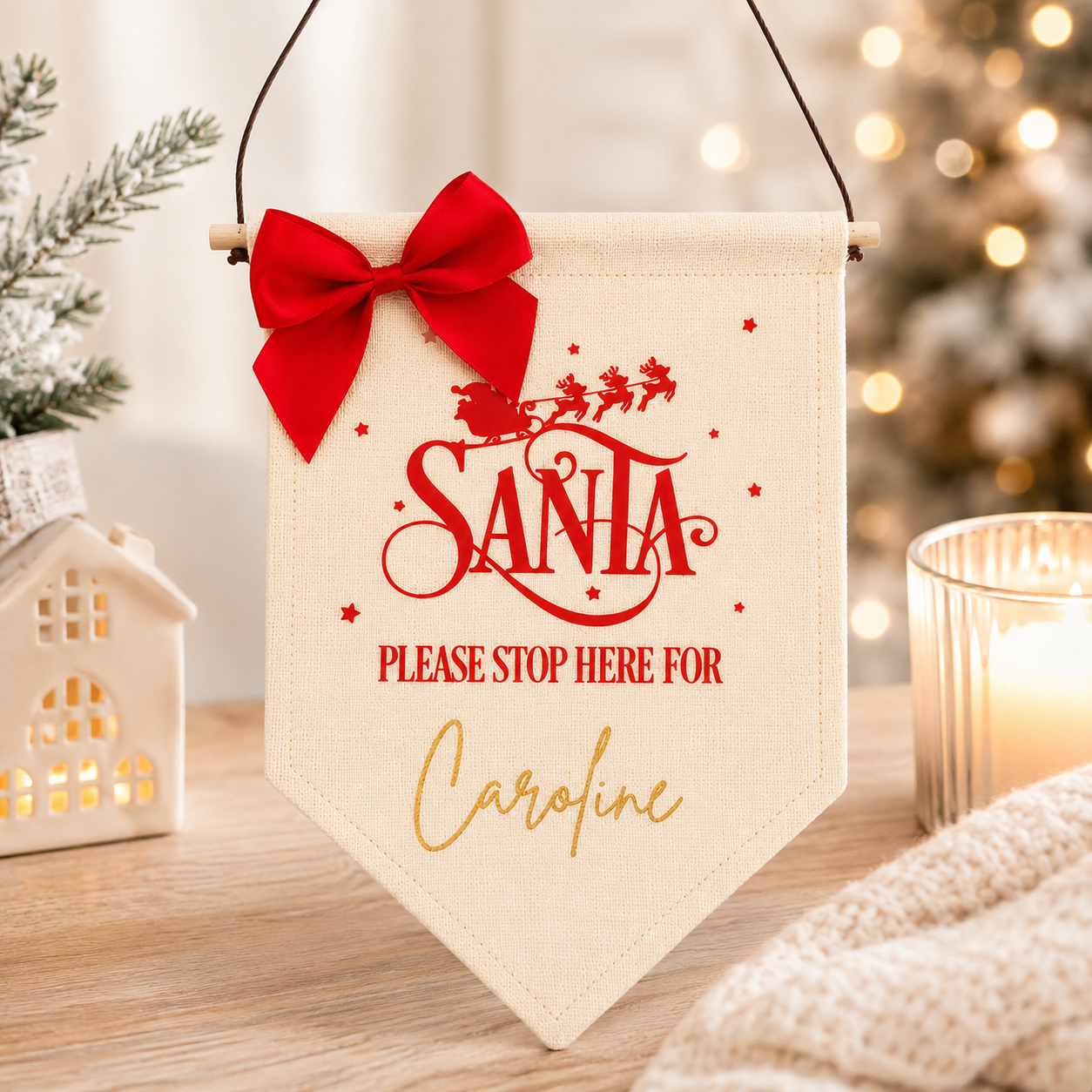 Decorative sign with 'Santa Please Stop Here For Caroline' text, red bow, and Christmas lights in the background.