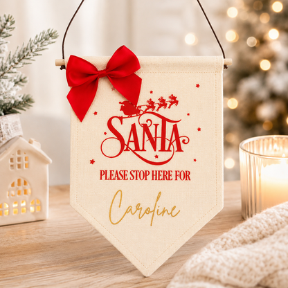 Decorative sign with 'Santa Please Stop Here For Caroline' text, red bow, and Christmas lights in the background.