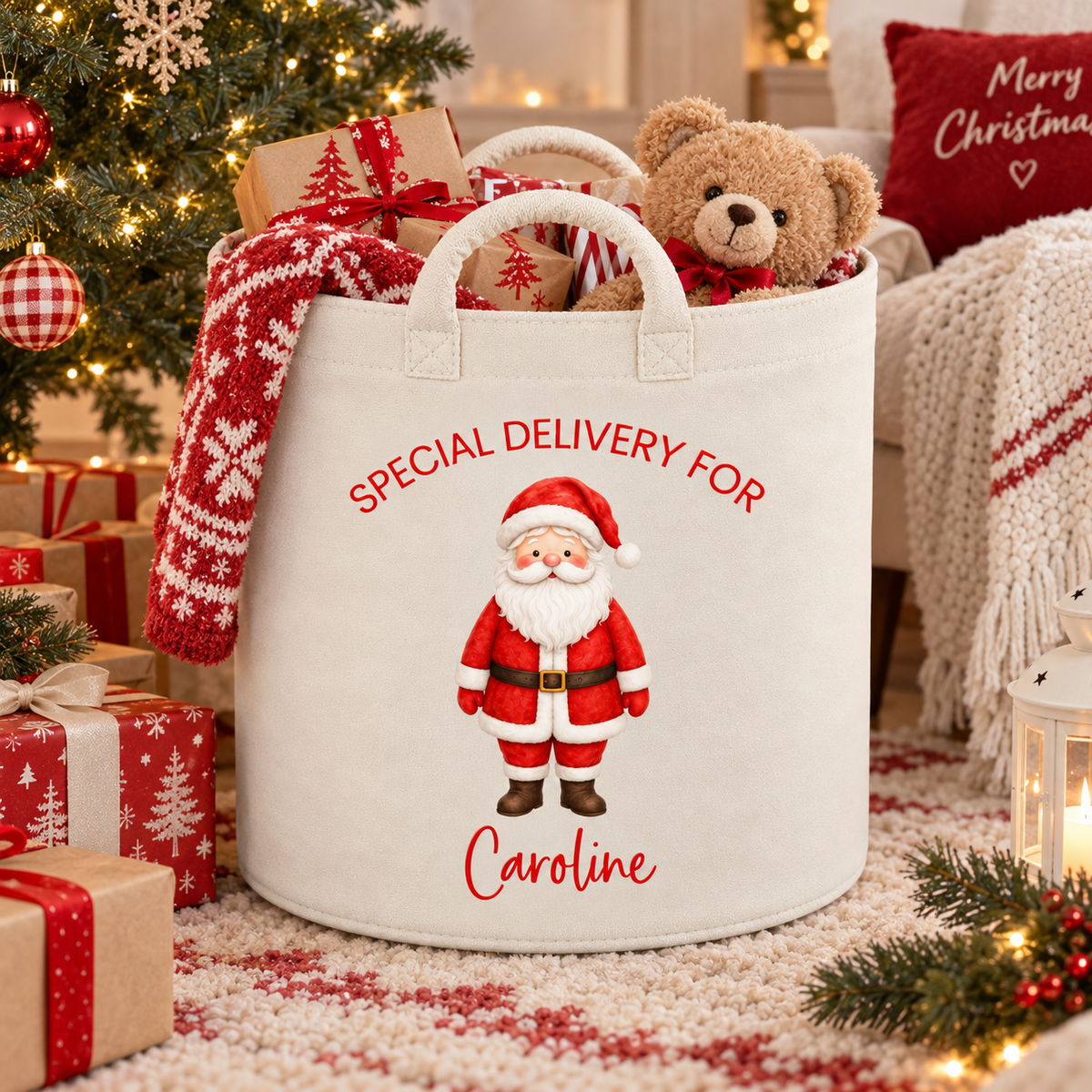 A white fabric storage basket with a personalized Christmas design, featuring an illustration of Santa Claus and the text 'SPECIAL DELIVERY FOR Caroline'. The basket is filled with holiday treats, a teddy bear, and a red knit blanket, set in a cozy, decorated living room next to a Christmas tree.