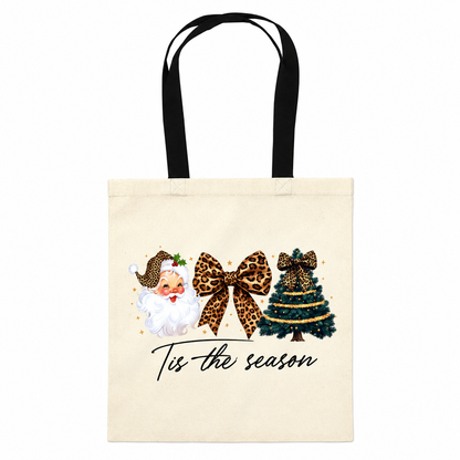 A cream canvas tote bag with black shoulder straps, featuring a festive Christmas graphic with a smiling Santa in leopard-print clothing, a large leopard-print bow, a decorated Christmas tree, and the text 'Tis the season' in black script.