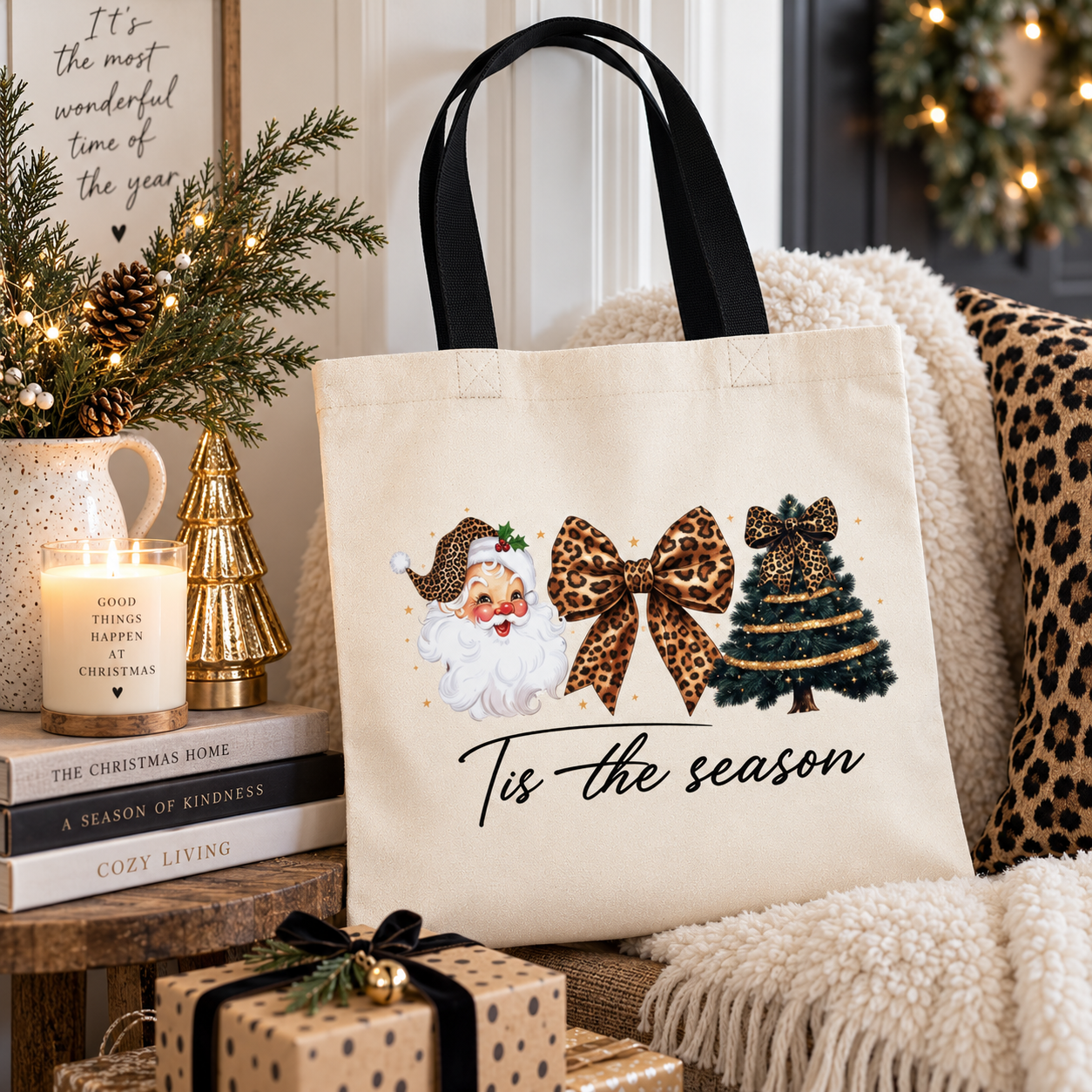 A cream canvas tote bag with black handles sits on a cozy chair in a decorated living room. The bag features a festive graphic of Santa Claus, a leopard-print bow, a Christmas tree, and the text 'Tis the season'.